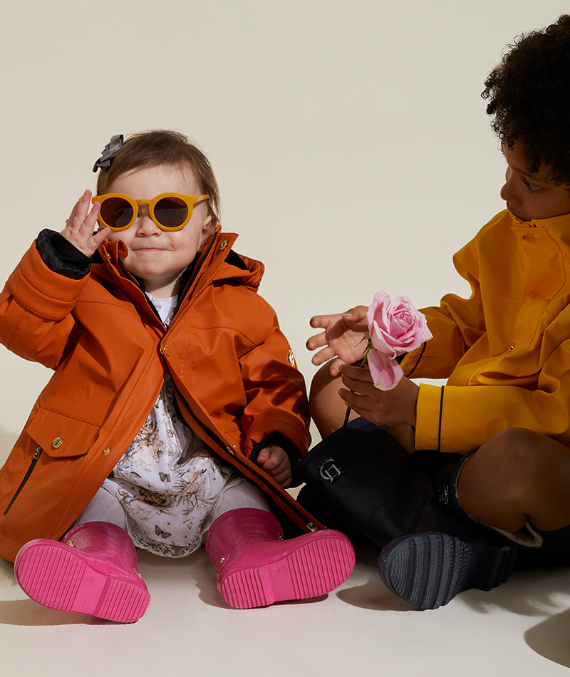 Little girl wears orange sunglasses with childrens rain jacket and floral dress with pink wellingtons sat next to little boy in yellow rain coat and navy blue wellingtons