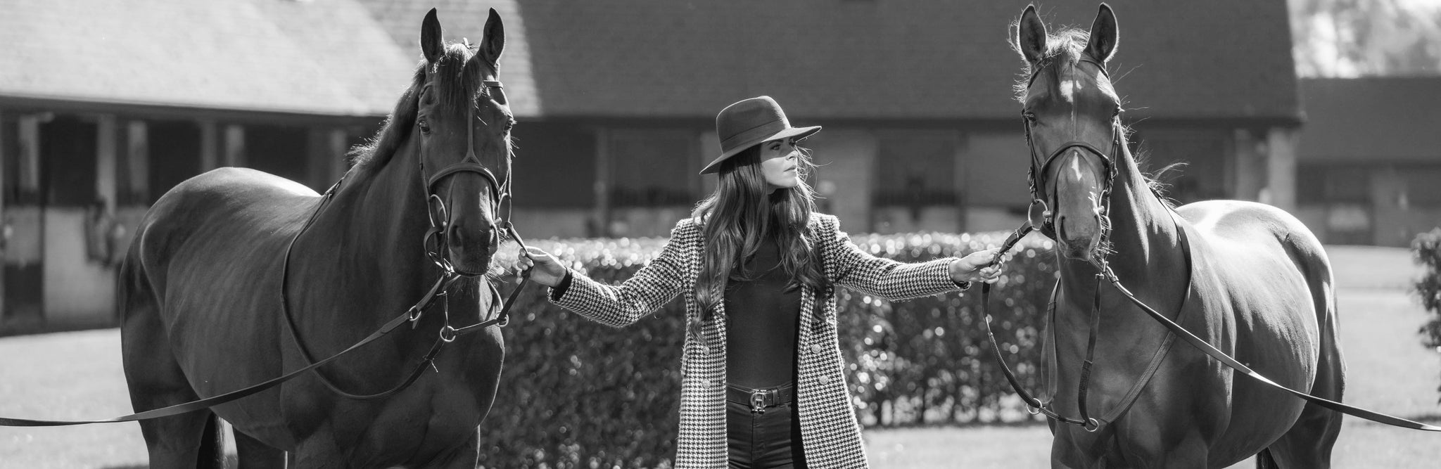 Black and white image with woman stood between two horses holding the bridle dressed in trilby hat and long houndstooth coat
