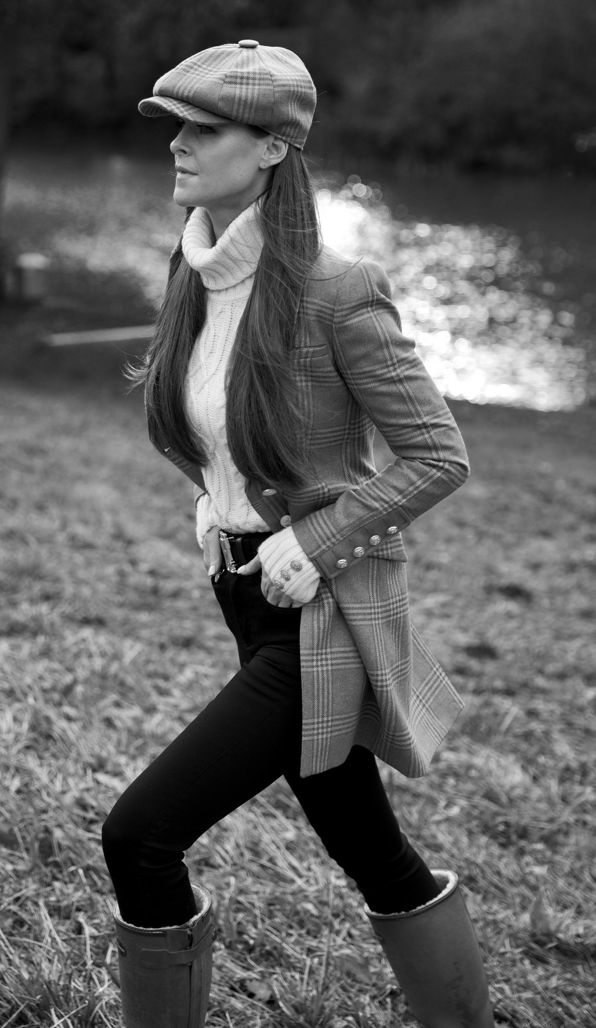 Black and white image of Holland Cooper owner, Jade Holland Cooper wearing baker boy cap, tweed coat, roll neck cable knit jumper, skinny jeans and sherpa lined wellingtons