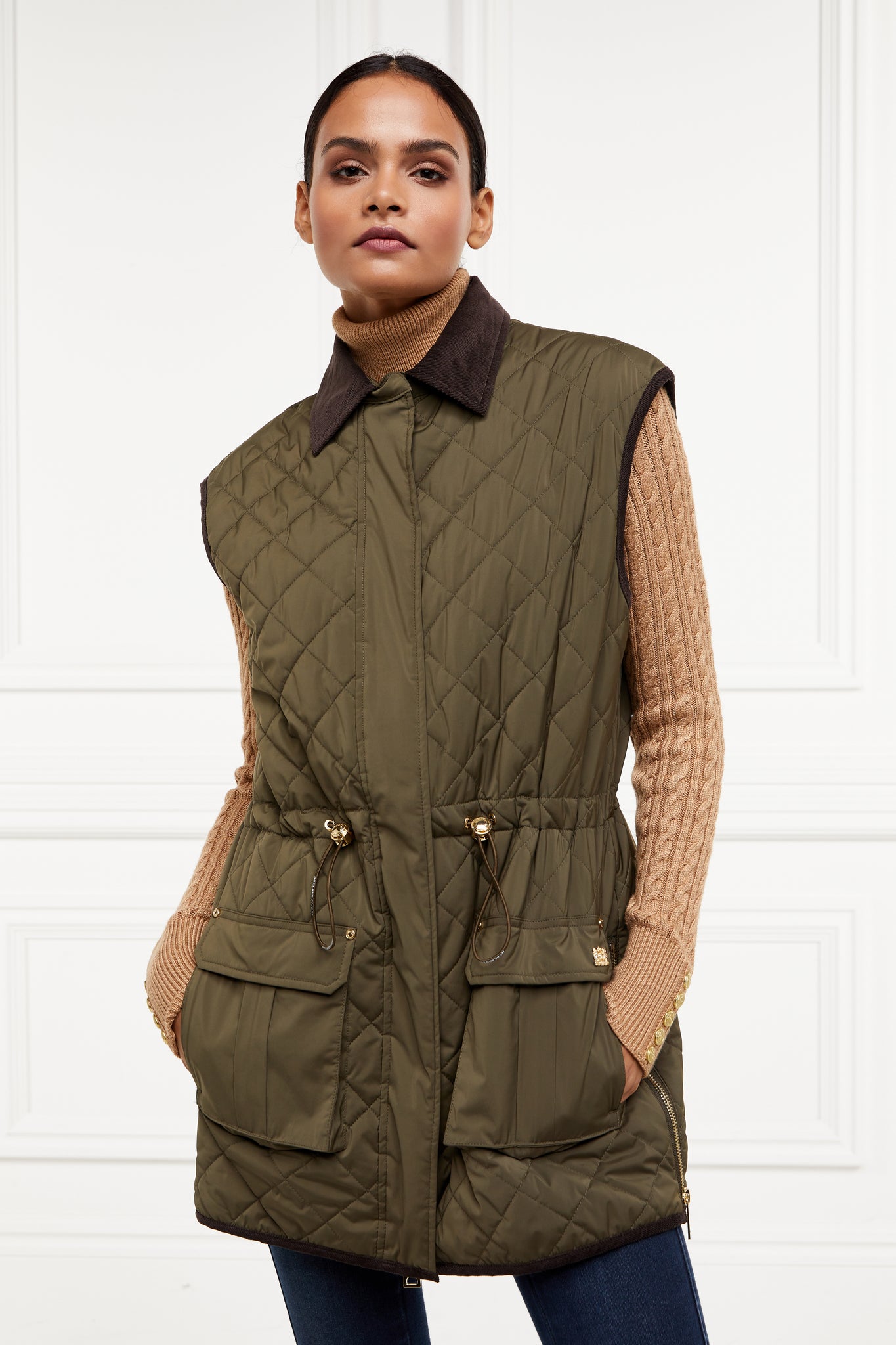 womens khaki quilted gilet with a dark brown collar and seams along the arm holes with drawstring toggles on the waist and two front pockets worn with a beige knitted jumper