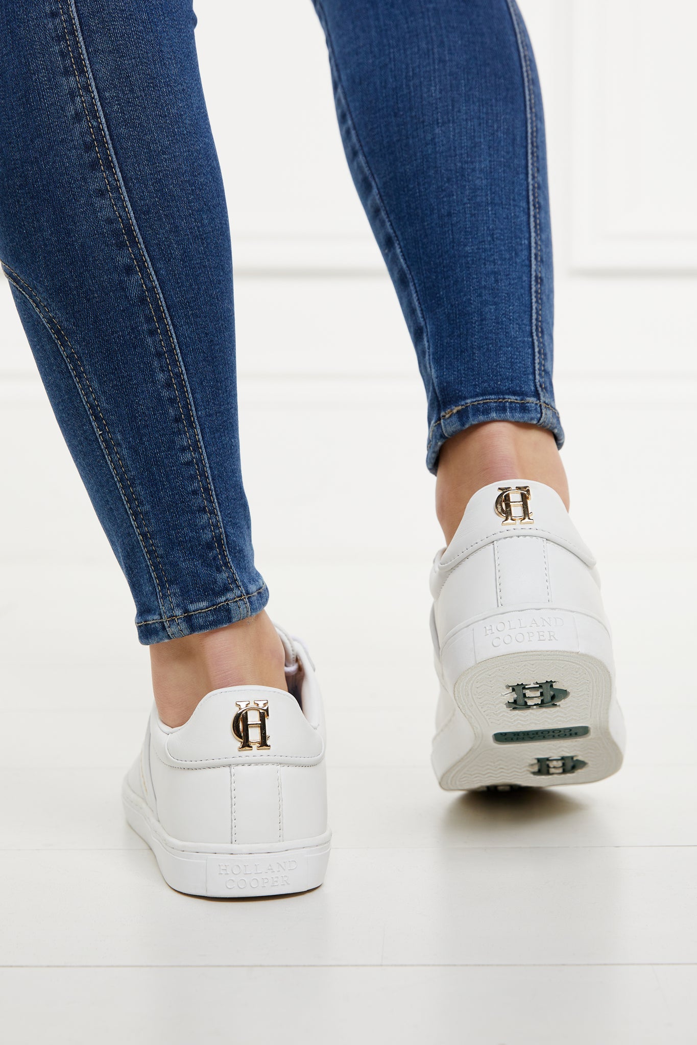 white leather trainers with white laces detailed with gold hardware, showing the bottom of the sole and the dark green branding. Worn with denim skinny jeans.