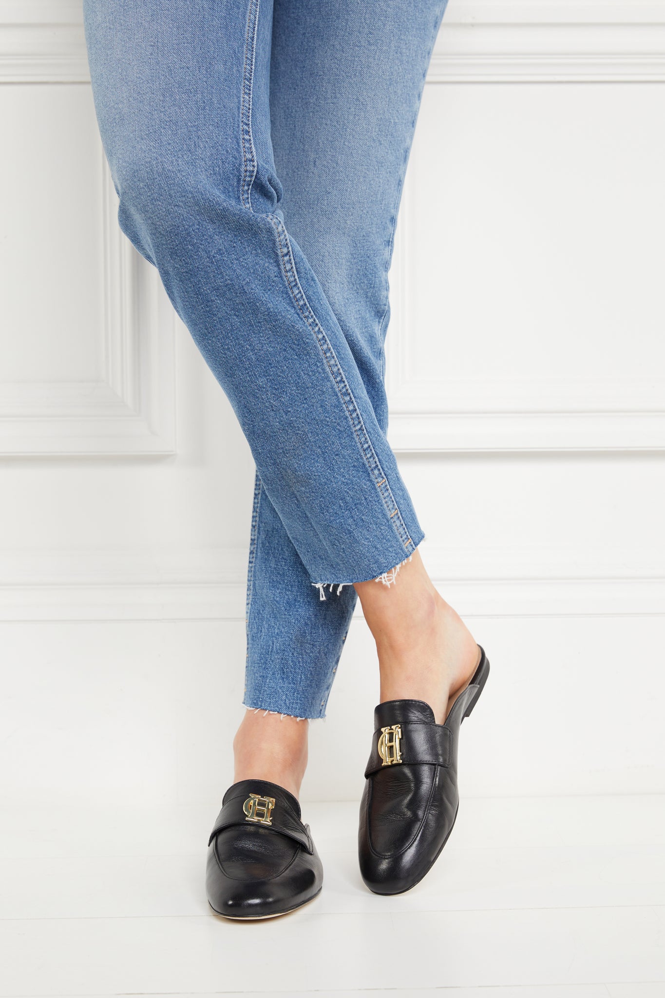 Close up of model wearing black leather backless loafers with a slightly pointed toe and gold hardware to the top paired with a pair of straight leg jeans with a frayed hem