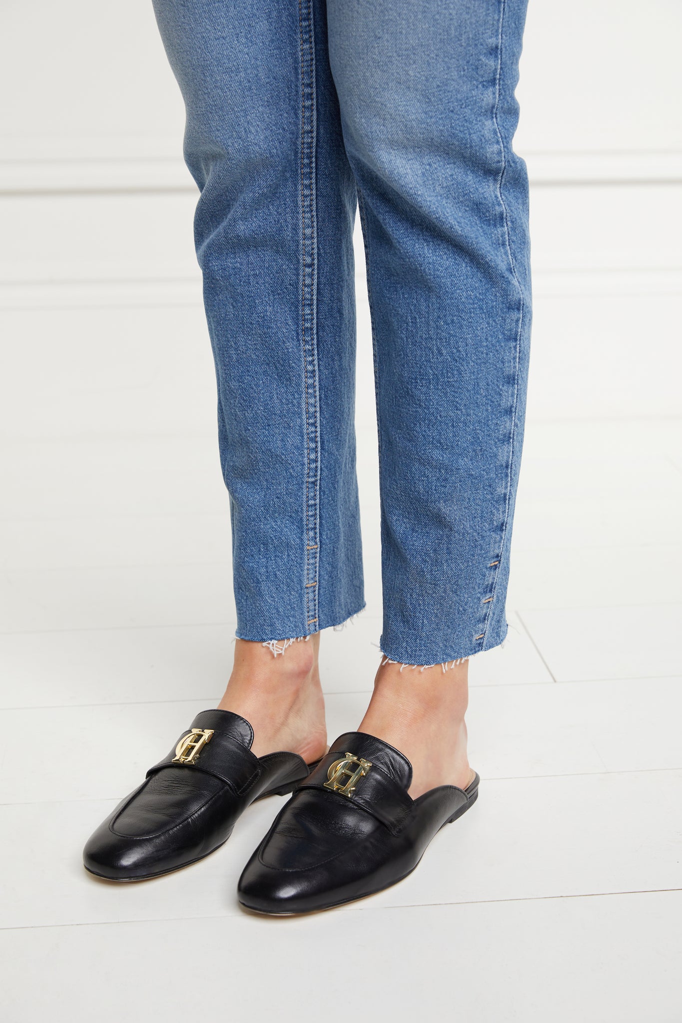 Side shot of model wearing black leather backless loafers with a slightly pointed toe and gold hardware to the top paired with a pair of straight leg jeans with a frayed hem