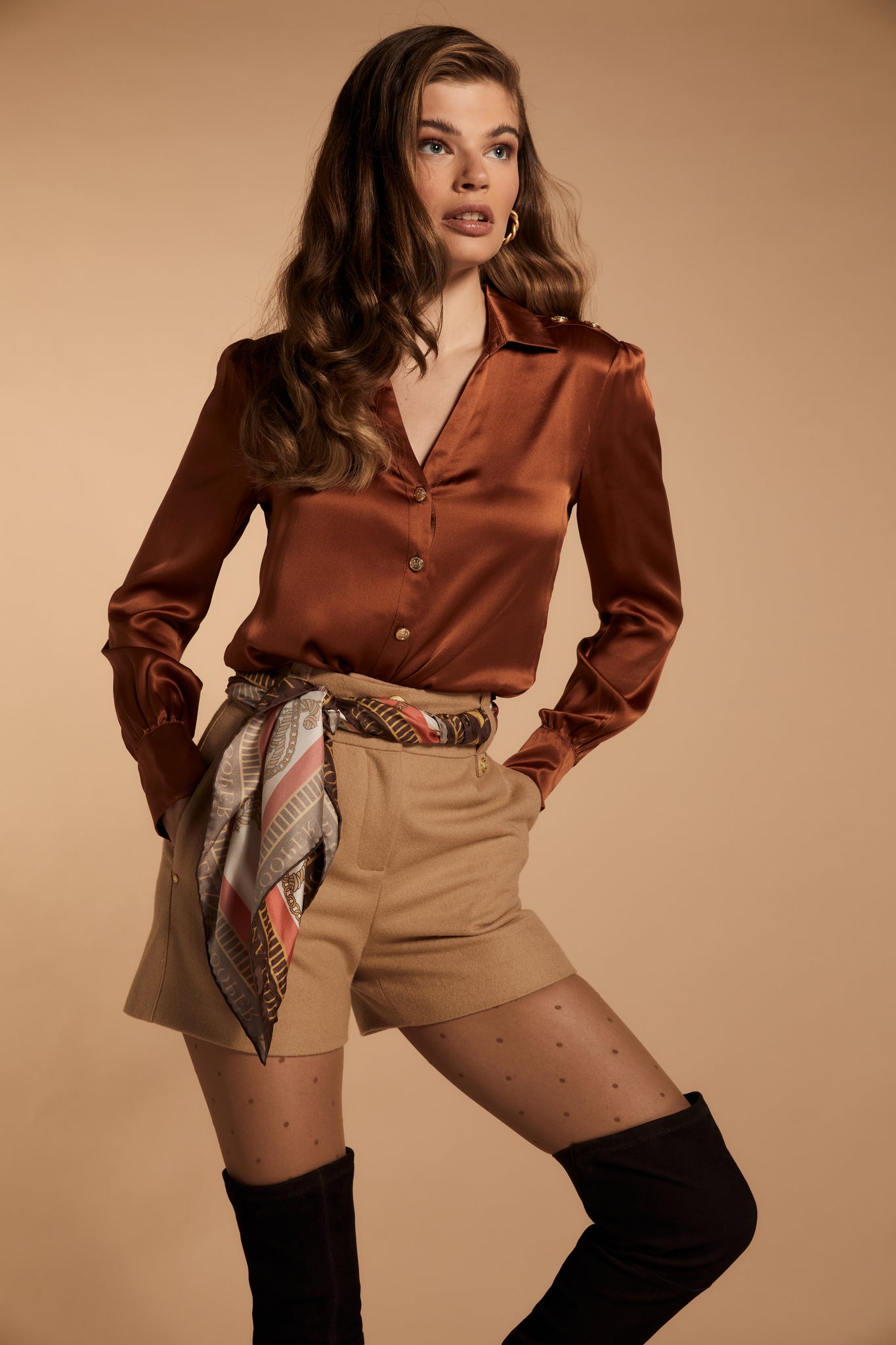 womens camel high rise tailored shorts with two single knife pleats and centre front zip fly fastening with twin branded gold stud buttons and side hip pockets with branded rivet detailing at top and bottom of pockets worn with caramel v neck silk shirt