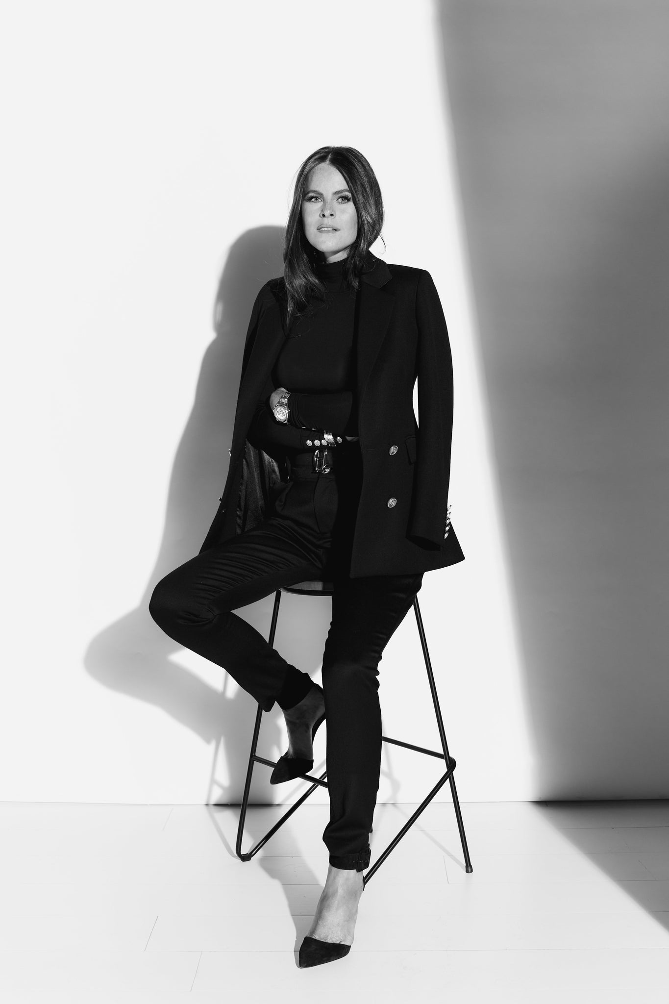 Black and white image of Jade Holland Cooper sat in studio on black metal stool dressed in womens black blazer, jumper, jeans and heels