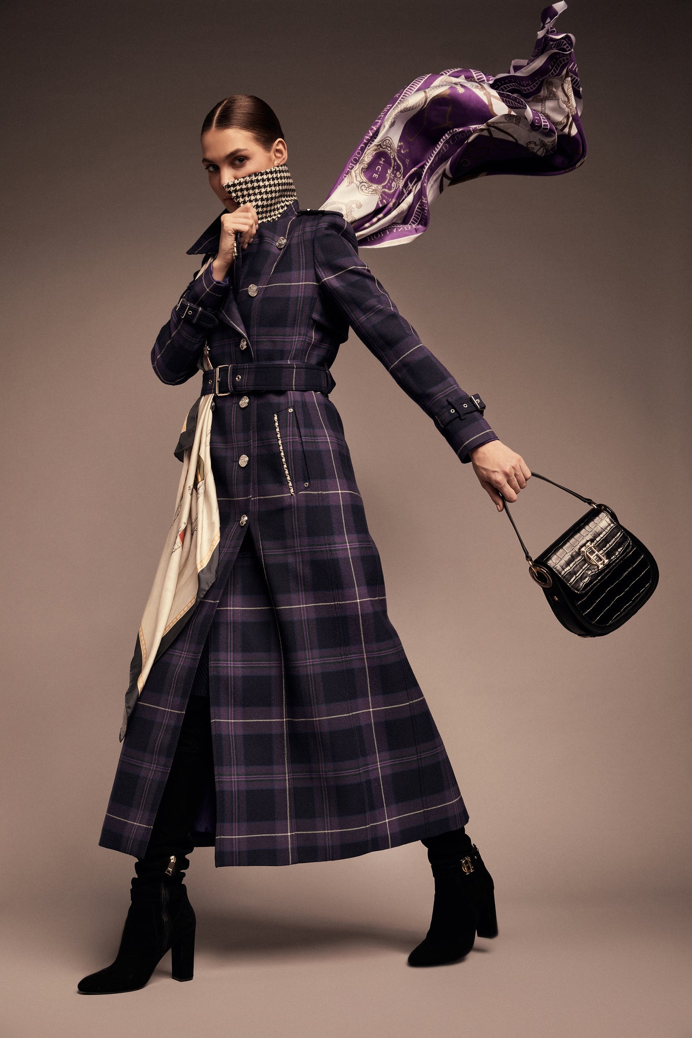 Model wearing purple tartan trench coat with silver buttons, flying purple silk scarf from neck and black croc saddle bag