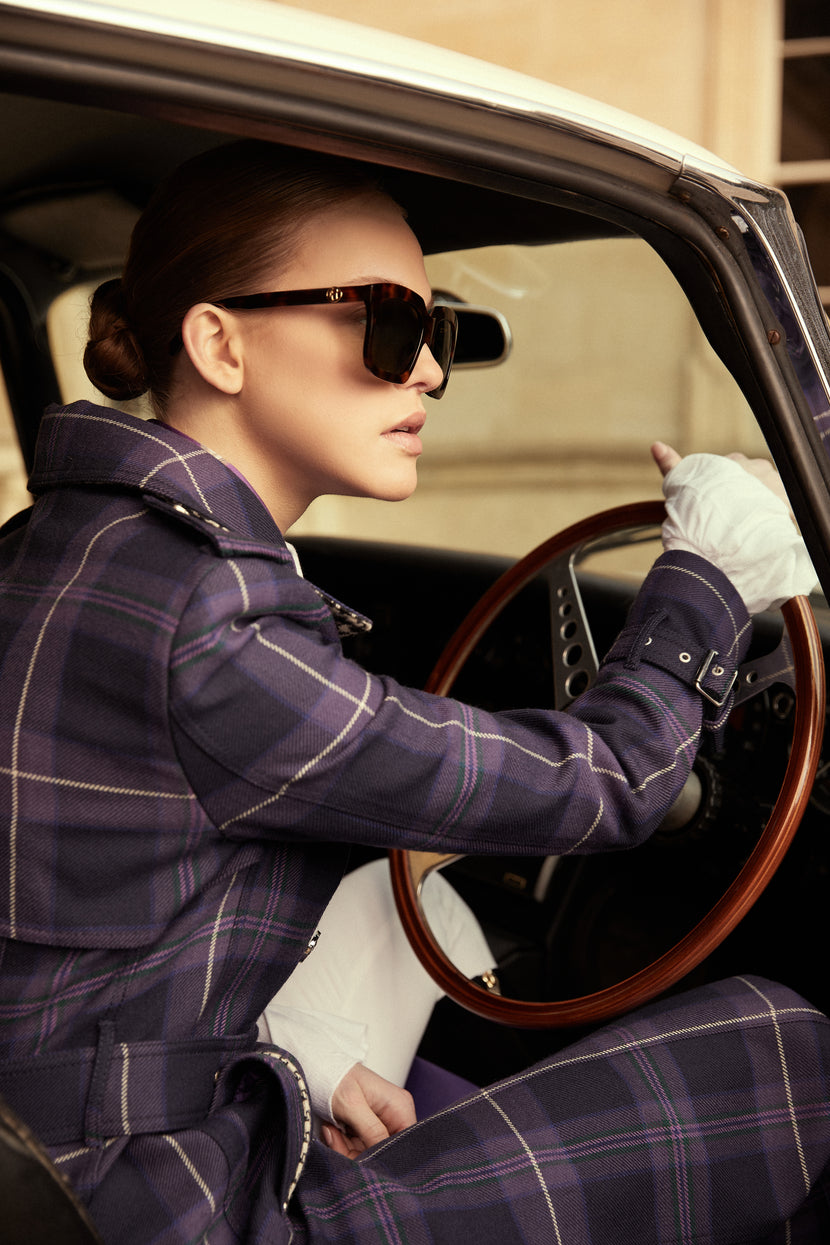 Woman sat in cream classic car in womens purple tartan trench coat with purple silk scarf and black sunglasses holding the wheel
