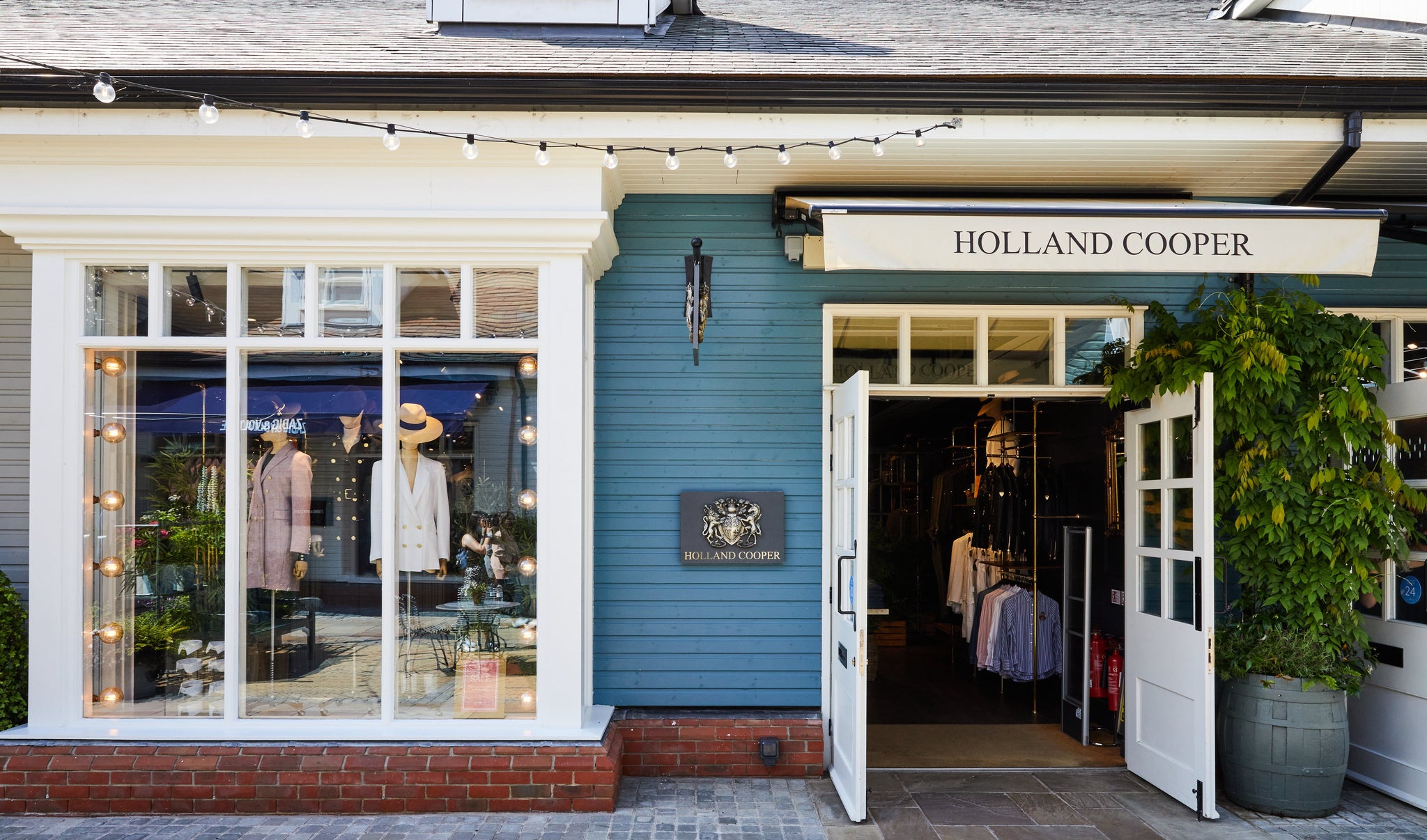 Holland Cooper Womens Bicester Store with black lettering and blue wooden building with white window and open glass door