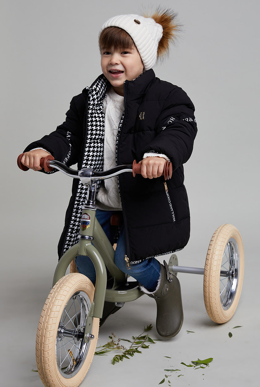 Little boy on green trike wearing white bobble hat with black puffer coat and green wellington's with blue jeans