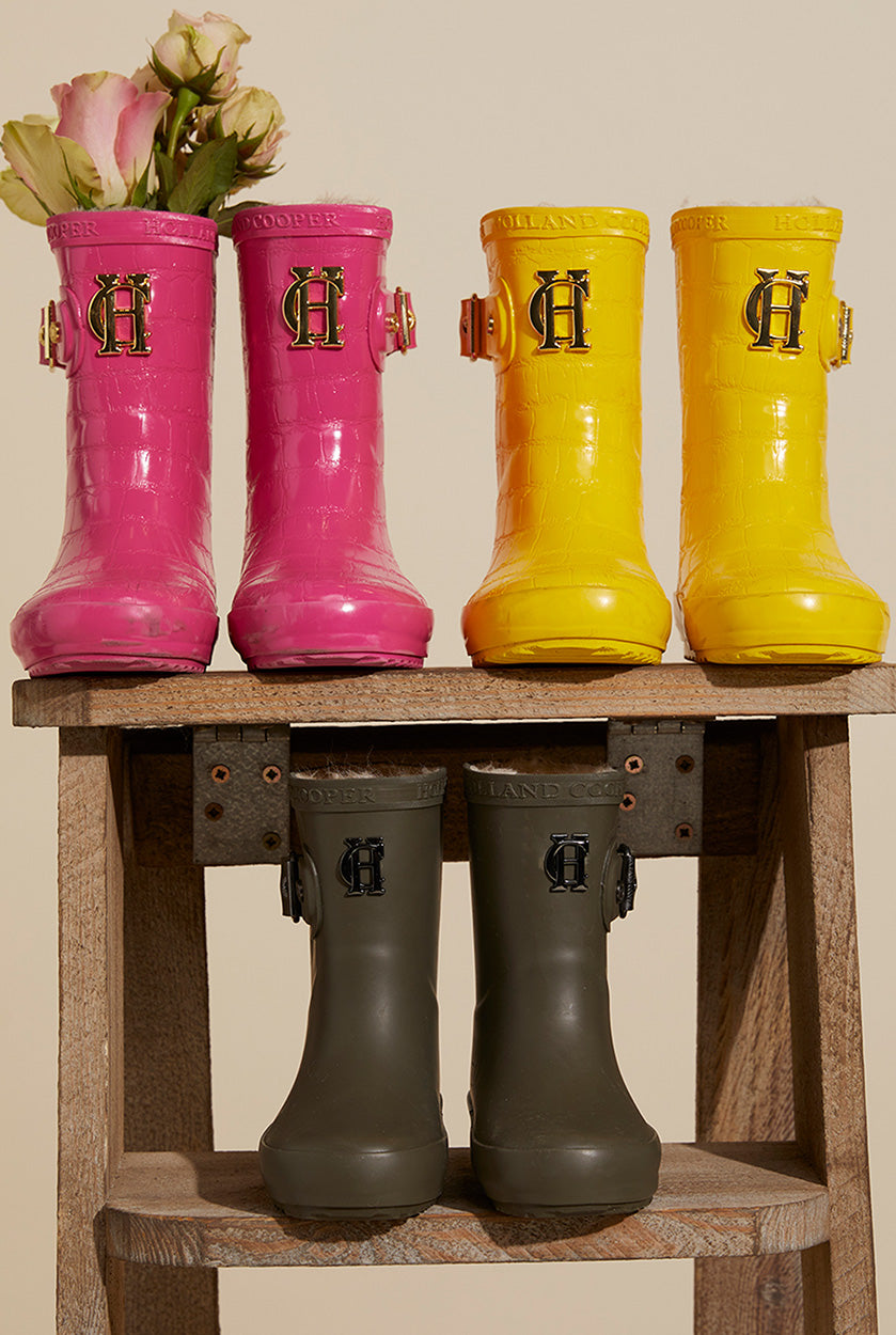 Three children's wellingtons stood on a wooden step in pink, yellow and green with flowers in them