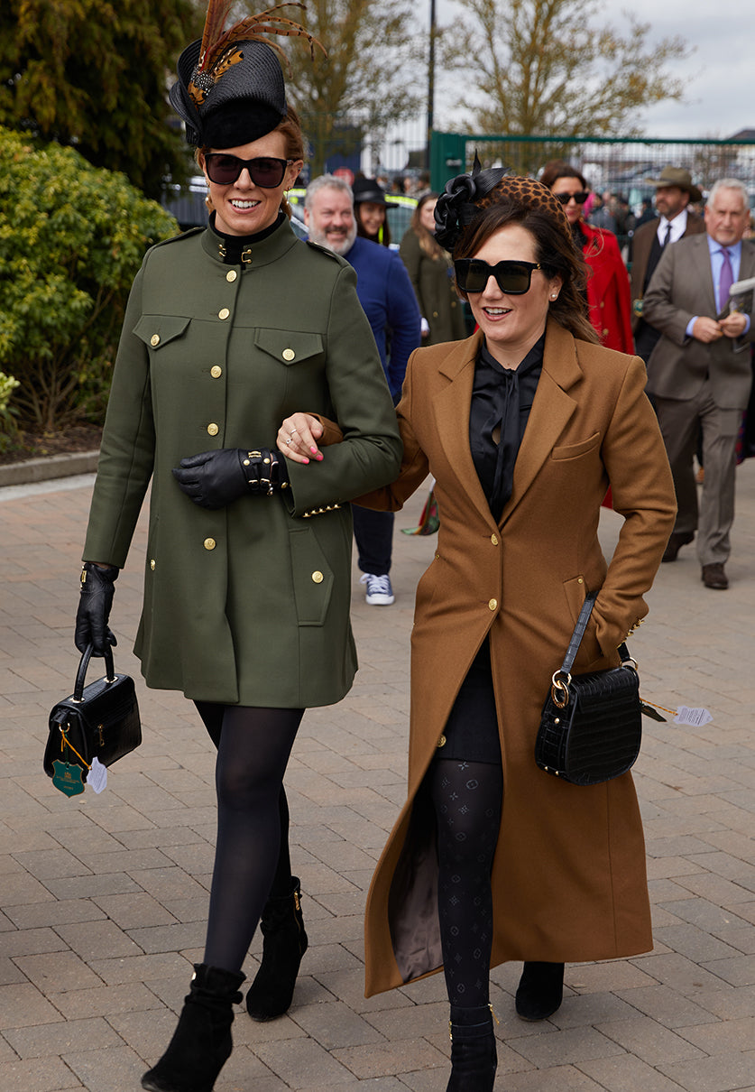 Two women walk arm in arm, one in khaki cape coat and black accessories and one in longline dark camel regency coat with black accessories