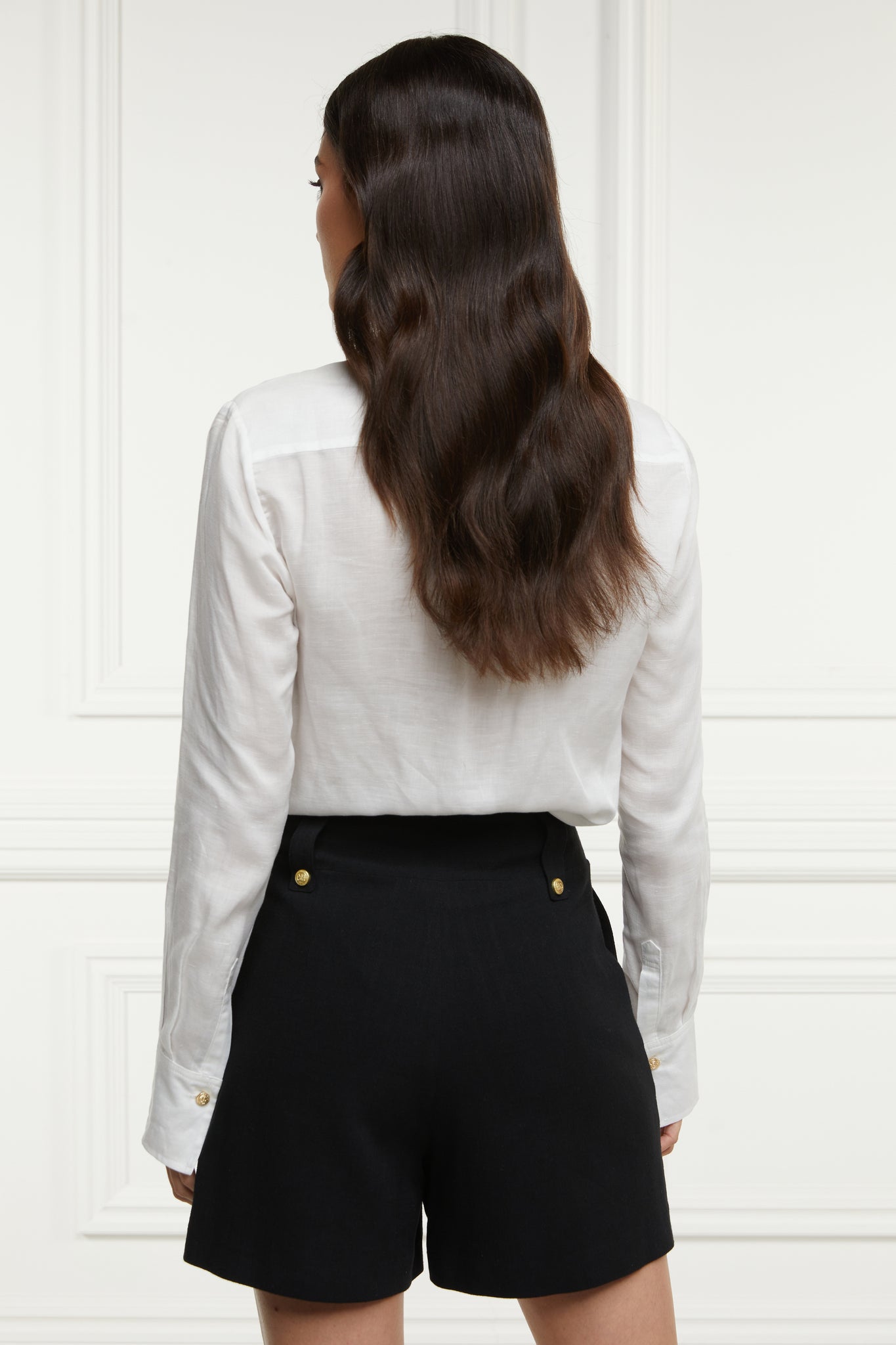 back of womens black linen tailored shorts with two single knife pleats and centre front zip fly fastening with two gold stud buttons worn with white classic shirt