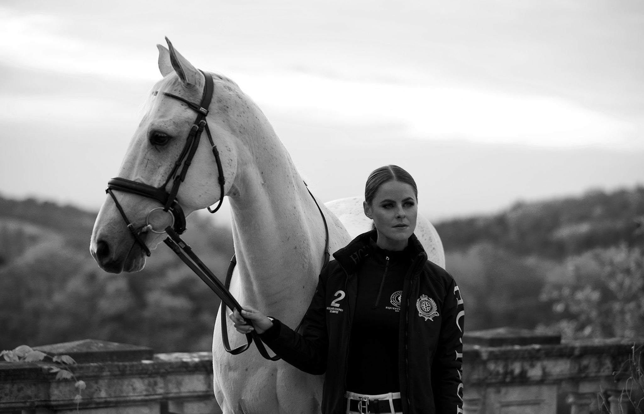 Black and white image of white horse held by the bridle by Jade Holland Cooper, owner of Holland Cooper wearing the team jacket with base layer