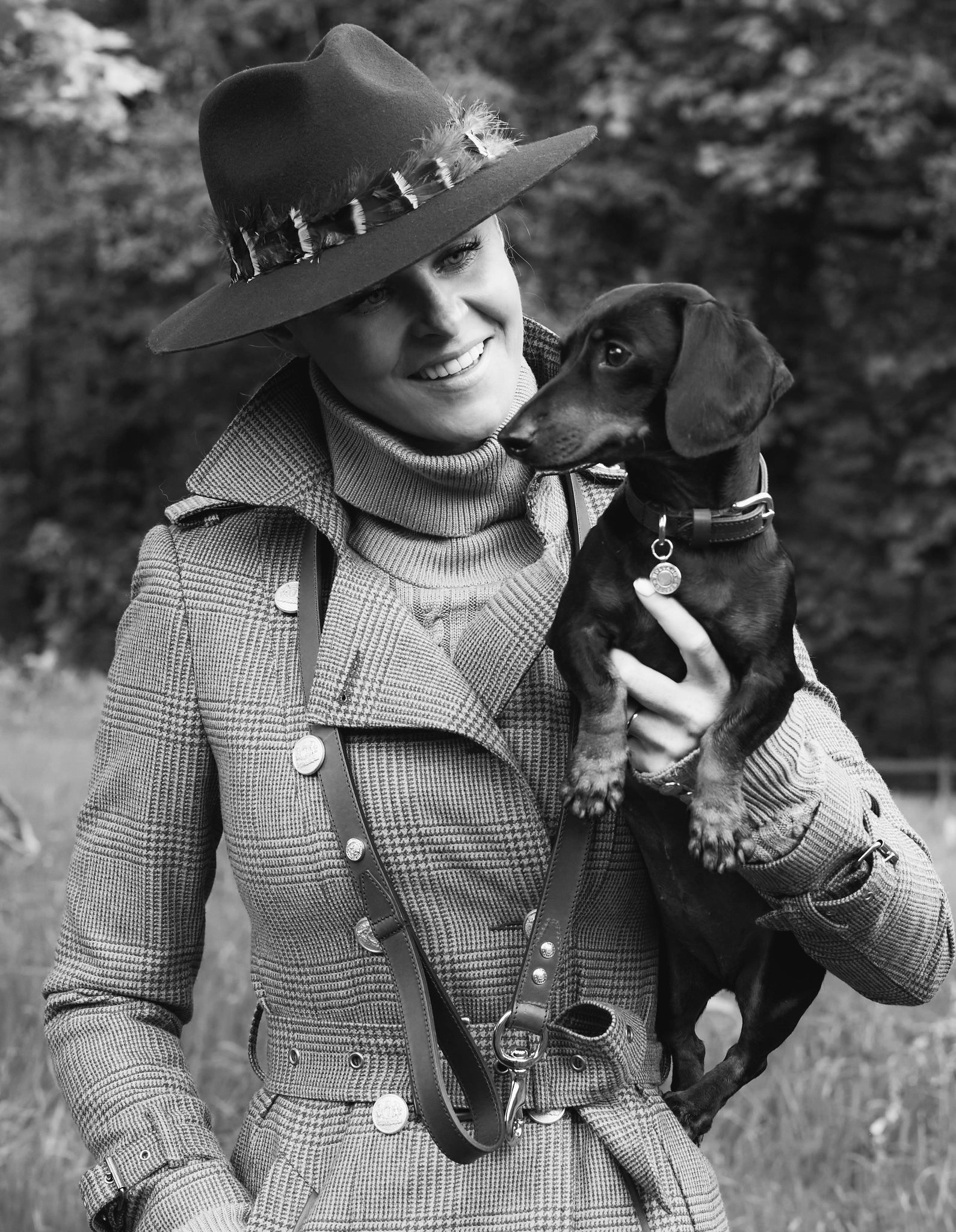 Jade Holland Cooper, owner of Holland Cooper wearing trilby hat, tweed trench coat with leather dog lead around neck holding miniature dachshund
