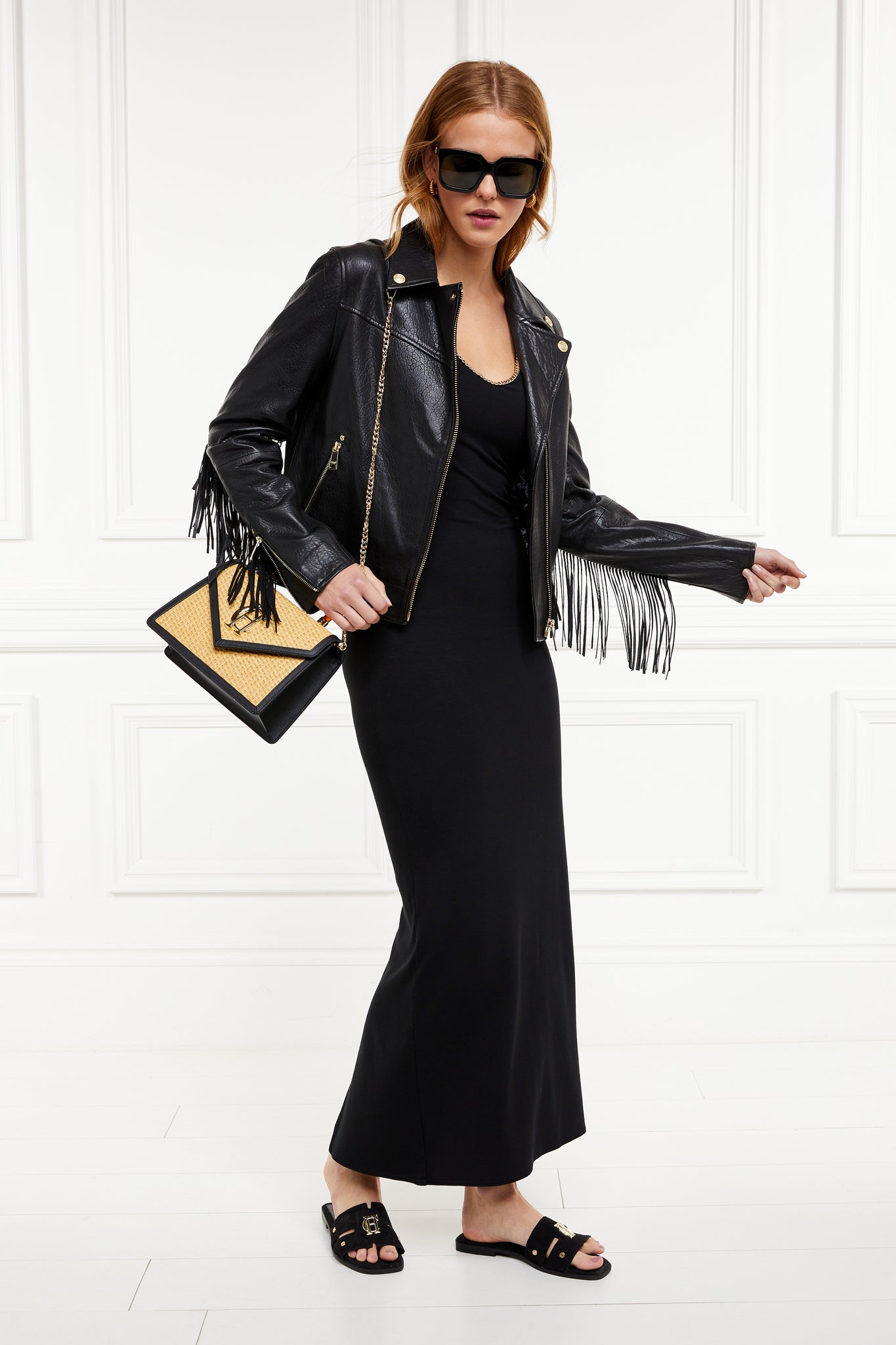 womens black v neck maxi dress with gold chain and black leather fringed biker jacket and black and natural raffia bag