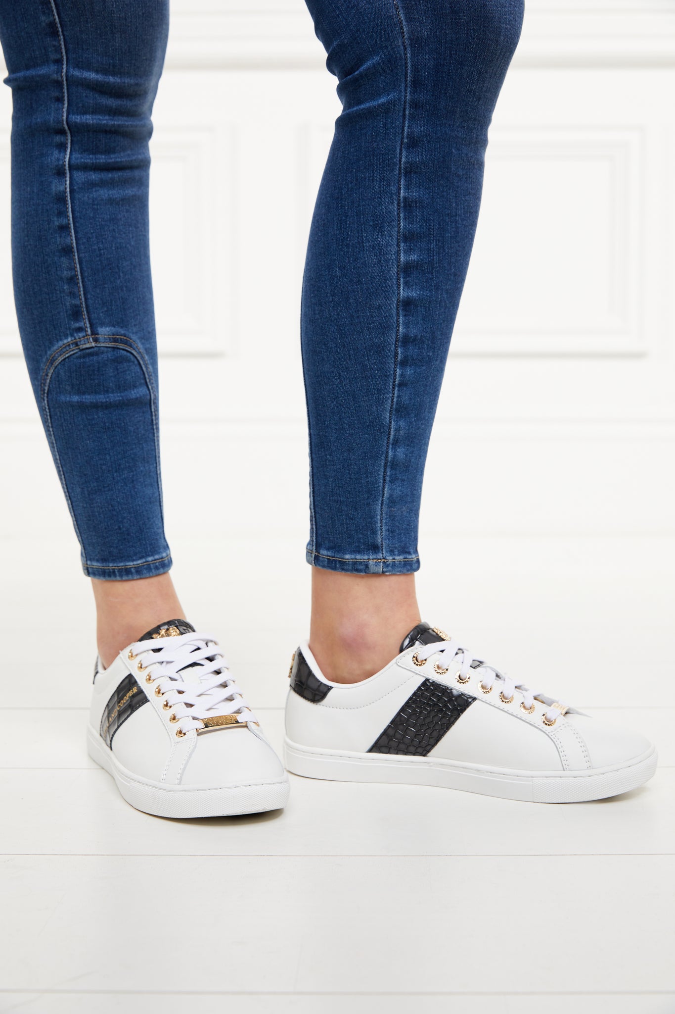 white leather trainers with white laces detailed with a diagonal stripe of black croc embossed leather with gold foil branding and a black croc embossed leather tongue and heel with gold hardware. Worn with skinny denim jeans.