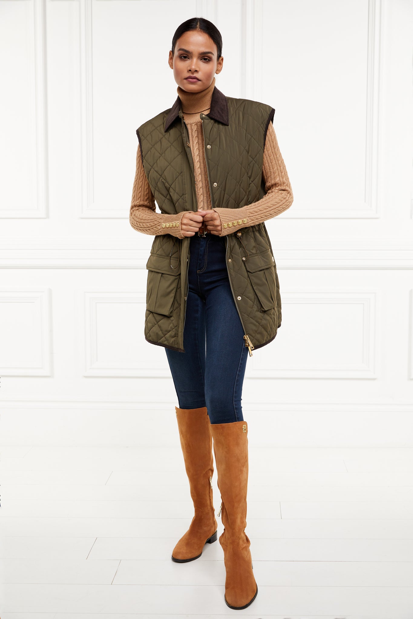  womens khaki quilted gilet with a dark brown collar and seams along the arm holes with drawstring toggles on the waist and two front pockets worn with a beige knitted jumper indigo skinny jeans and light brown suede knee high boots