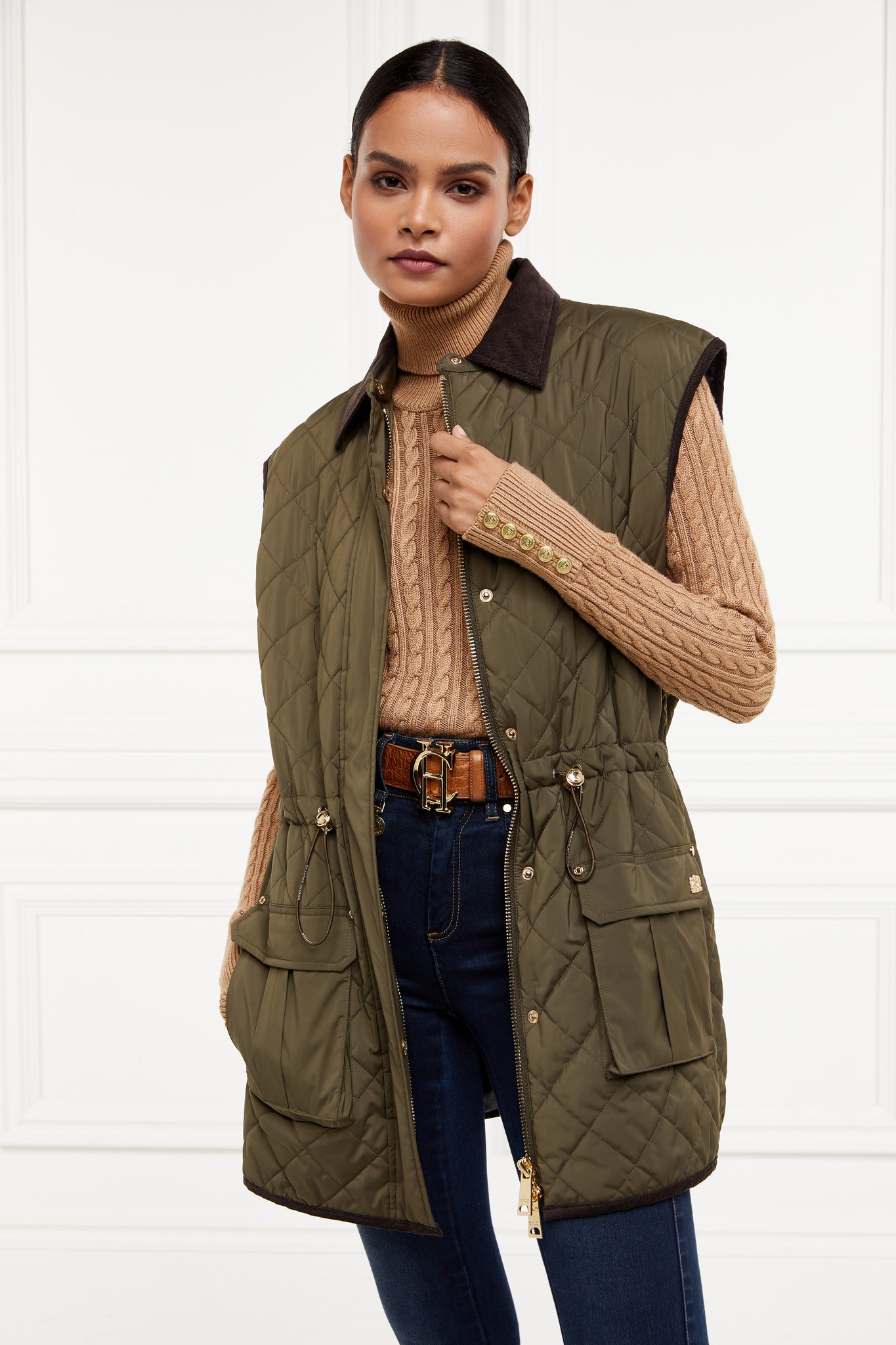 womens khaki quilted gilet with a black collar and seams along the arm holes with drawstring toggles on the waist and two front pockets worn with a beige knitted jumper a tan belt and indigo skinny jeans