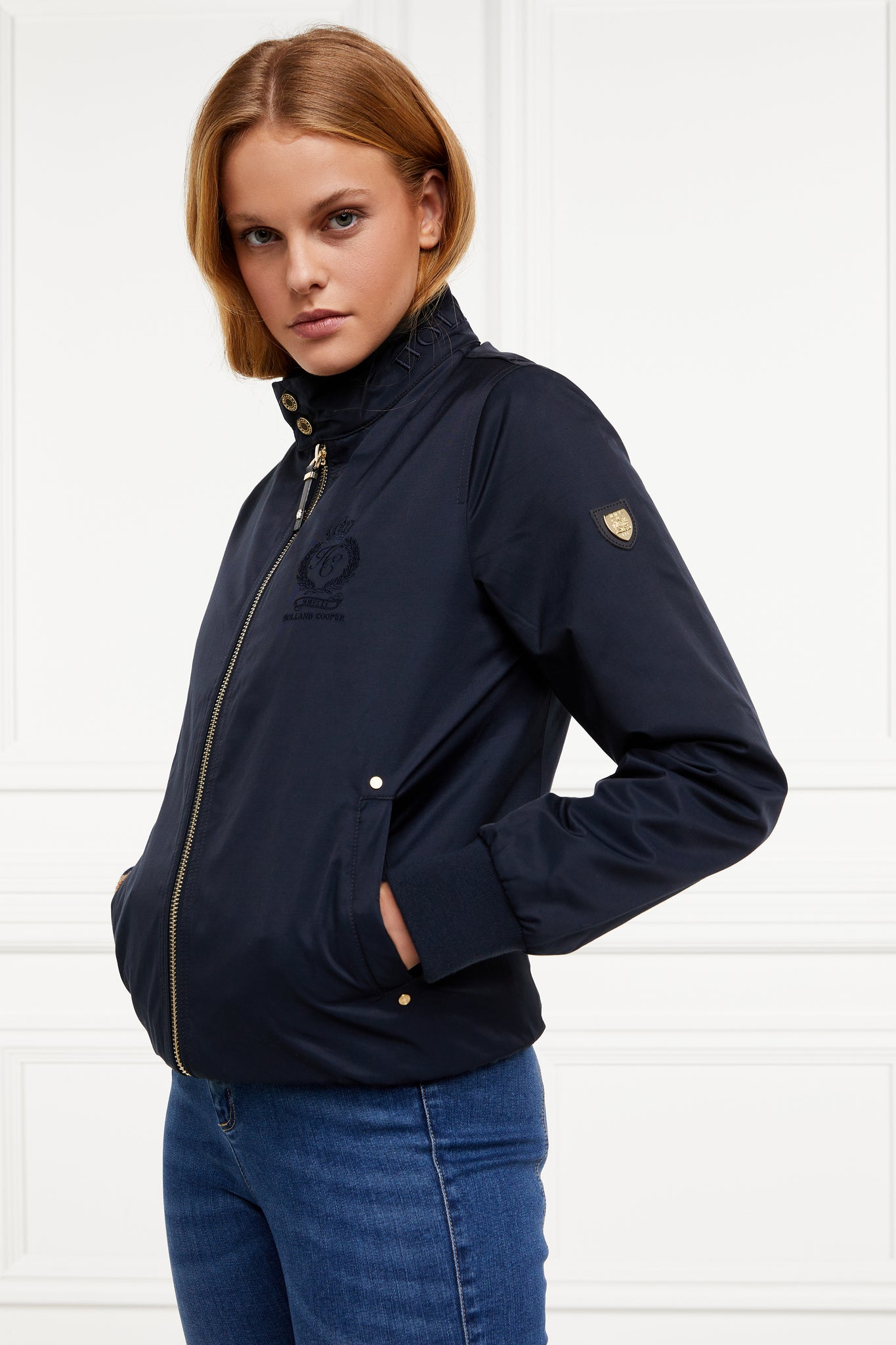 side of Womens navy 100% waterproof hoodless bomber style jacket with ribbed cuffs and hem, two pockets on hips a full zip down the centre front showing gold shield badge detail to top of arm