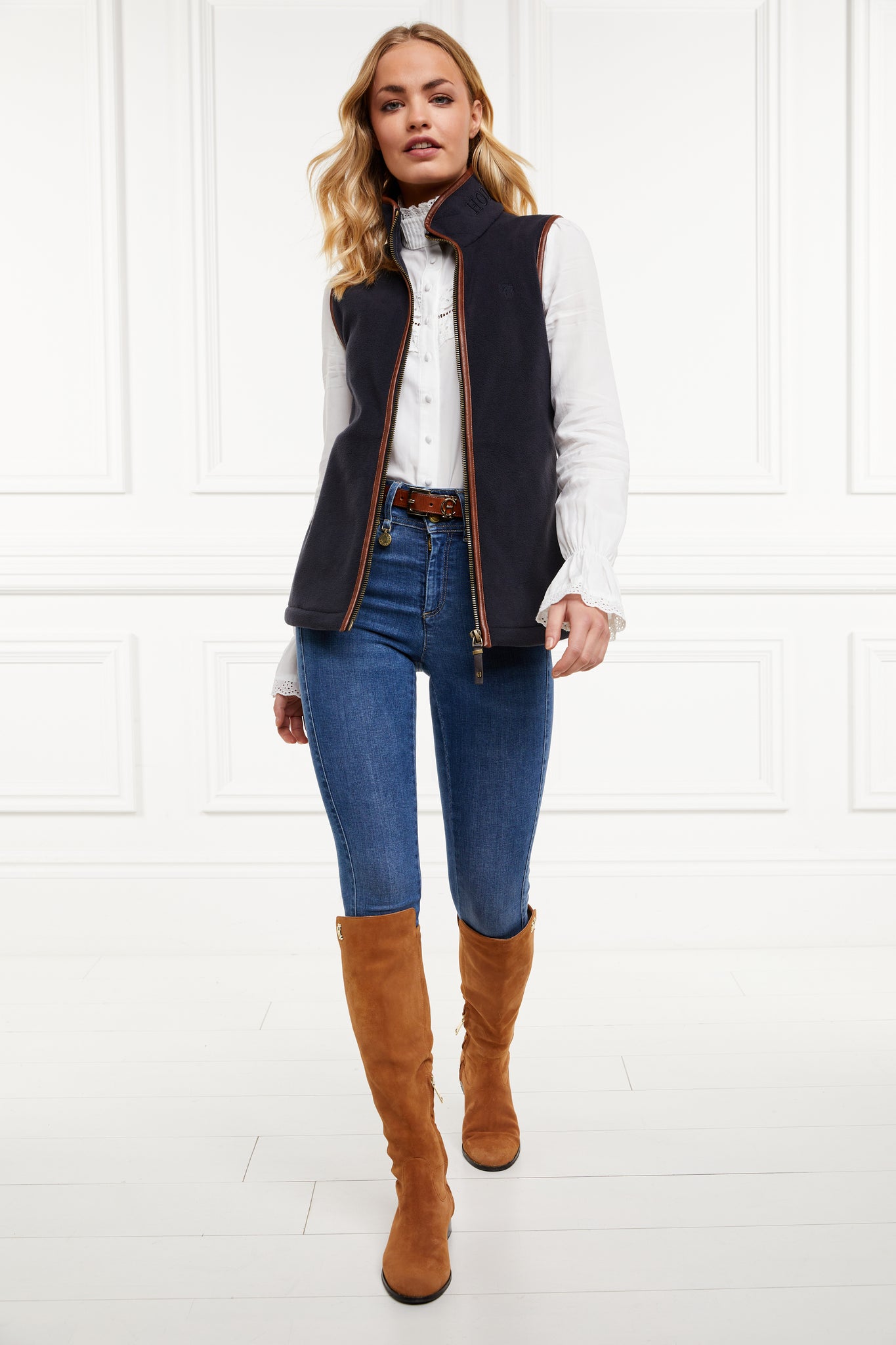 womens fleece gilet in navy with dark brown leather piping around armholes neckline and down the front zip fastening worn with a white shirt dark denim jeans a dark brown belt and tan suede knee high boots