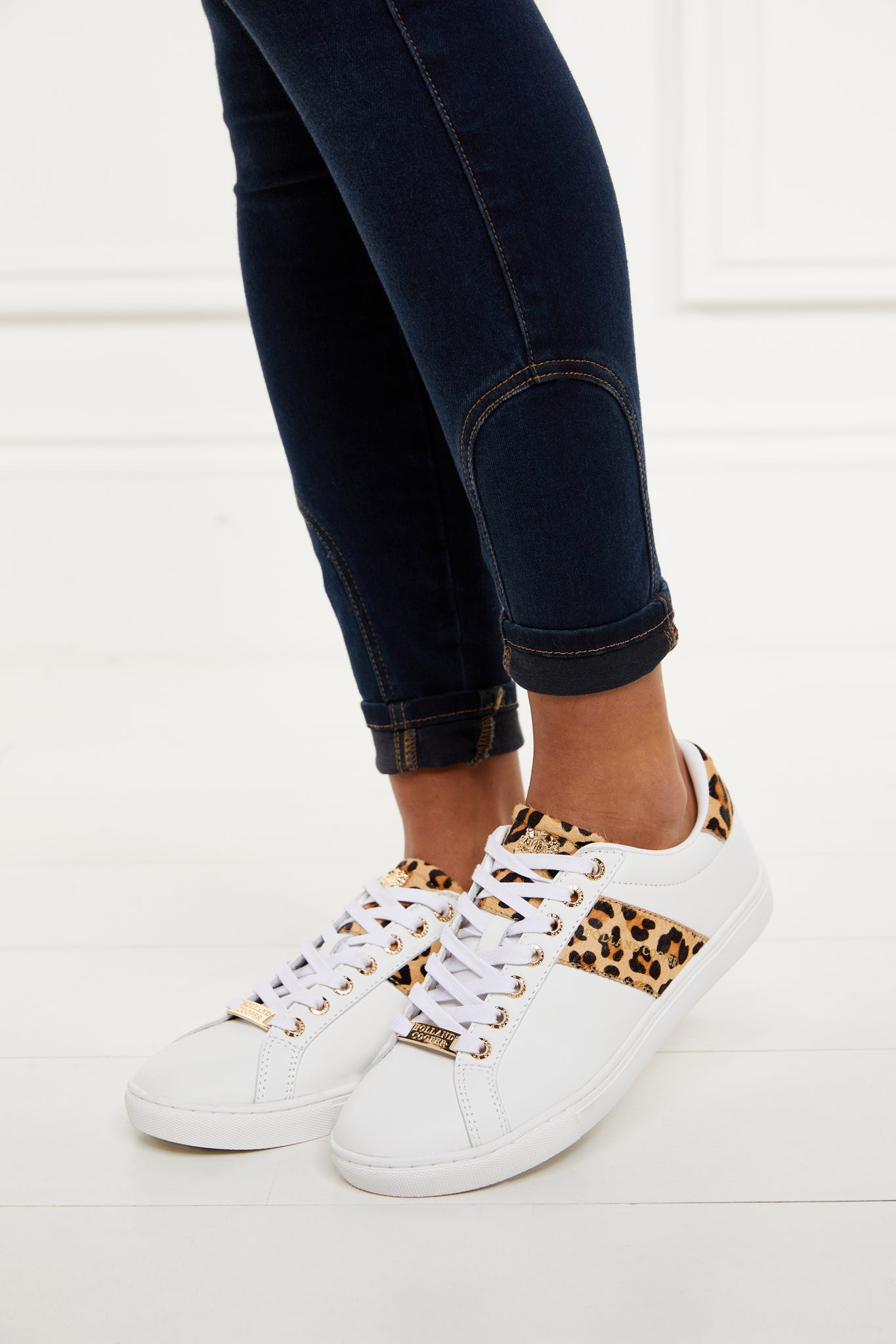 white leather trainers with white laces detailed with a diagonal stripe of leopard print on the side with gold foil branding and a leopard print heel and tongue with gold hardware. Worn with rolled up denim skinny jeans.