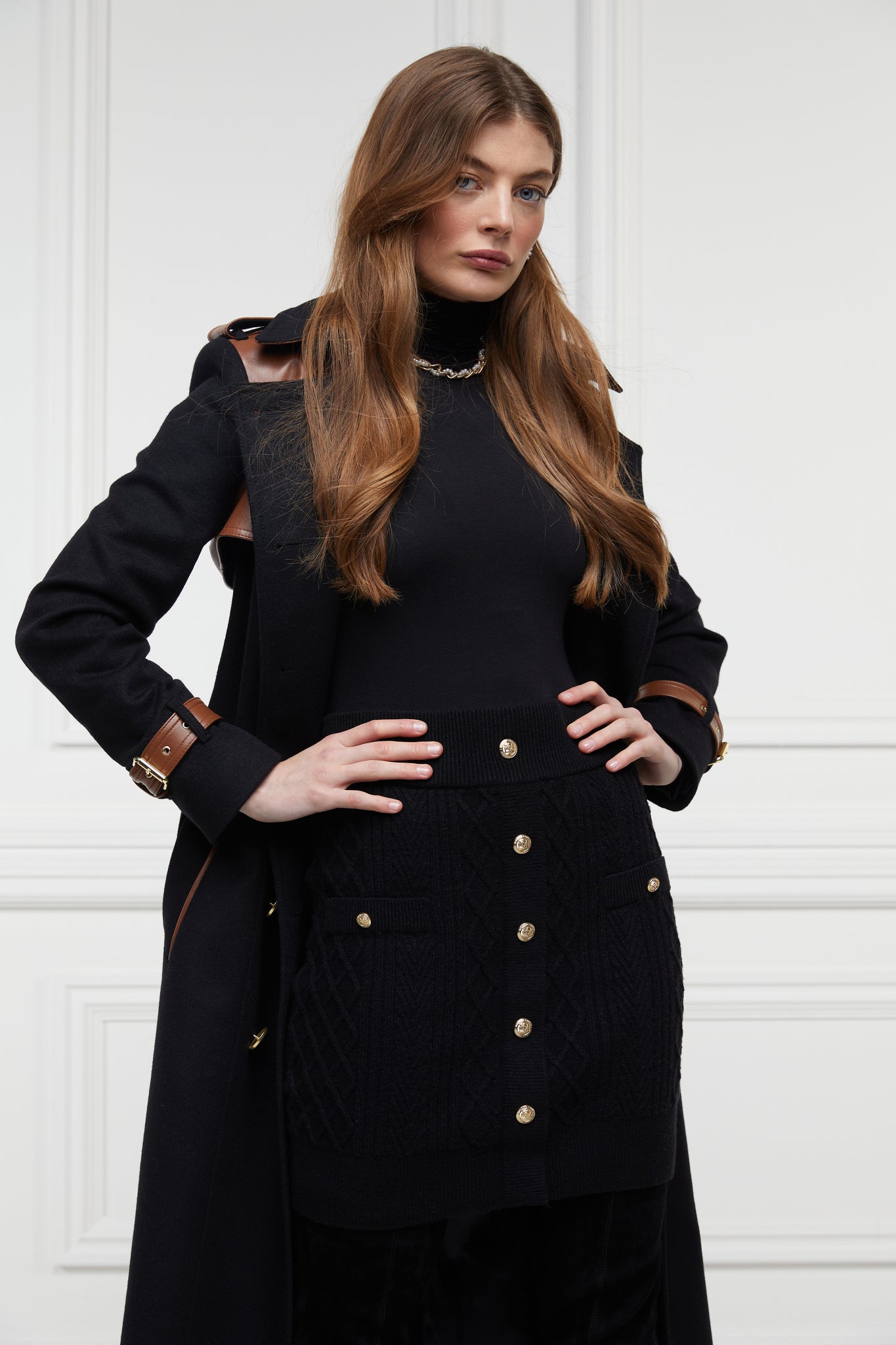 womens cable knit mini skirt with two welt pockets on each hip with gold button fastenings and gold button detailing down the centre front worn with ful length trench coat in black