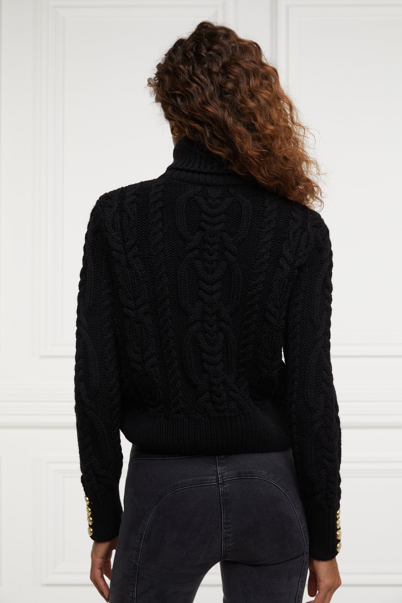 a chunky cable knit roll neck jumper in black with dropped shoulders and thick ribbed cable trims and gold buttons on cuffs and collar