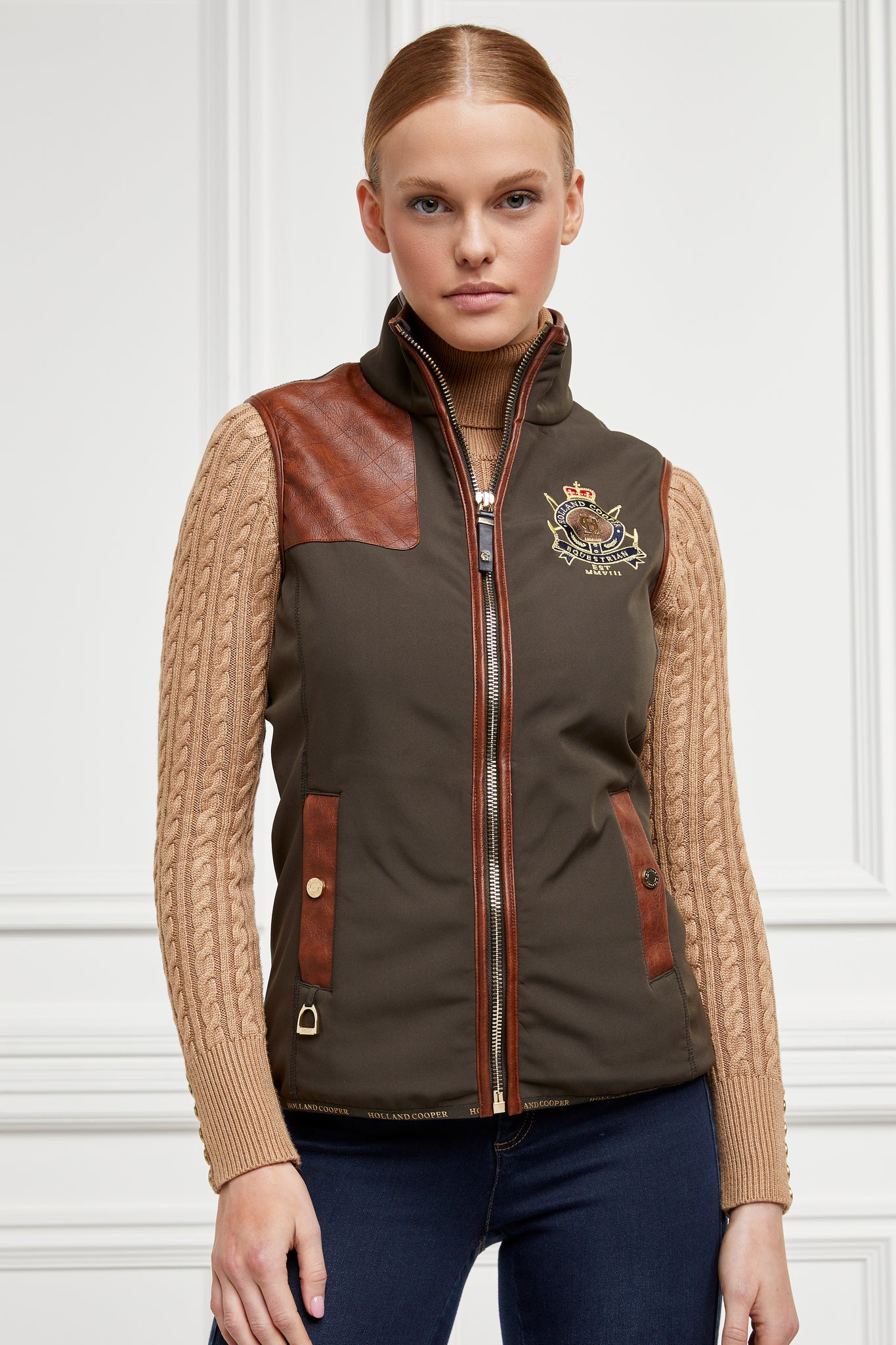 womens khaki gilet with dark brown leather seams along the arm holes pockets and down the zip with a gun patch on the shoulder and an embroidered logo worn with a beige knitted jumper and indigo jeans