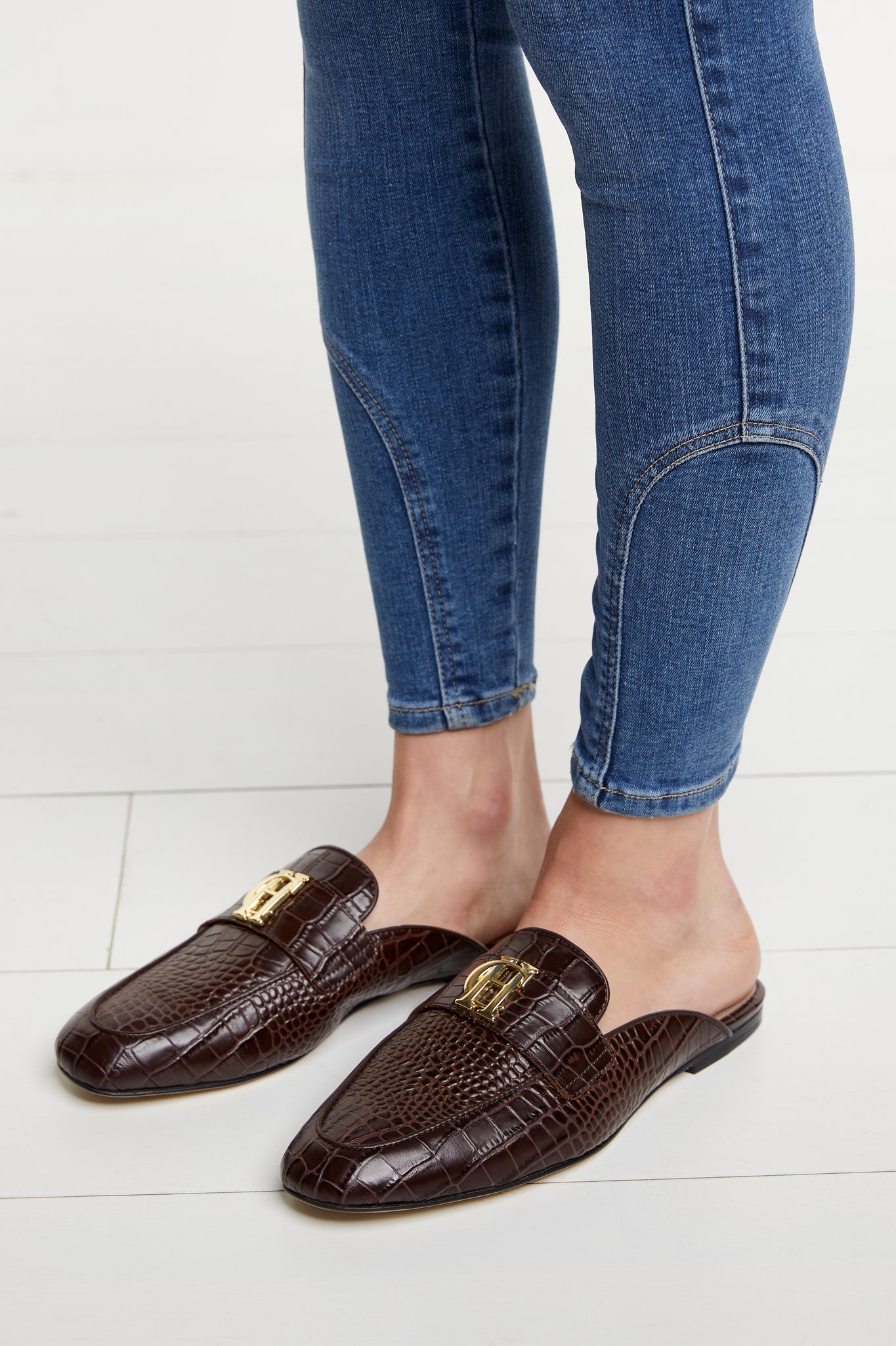 Side shot of brown croc embossed leather backless loafers with a slightly pointed toe and gold hardware to the top paired with denim skinny jeans