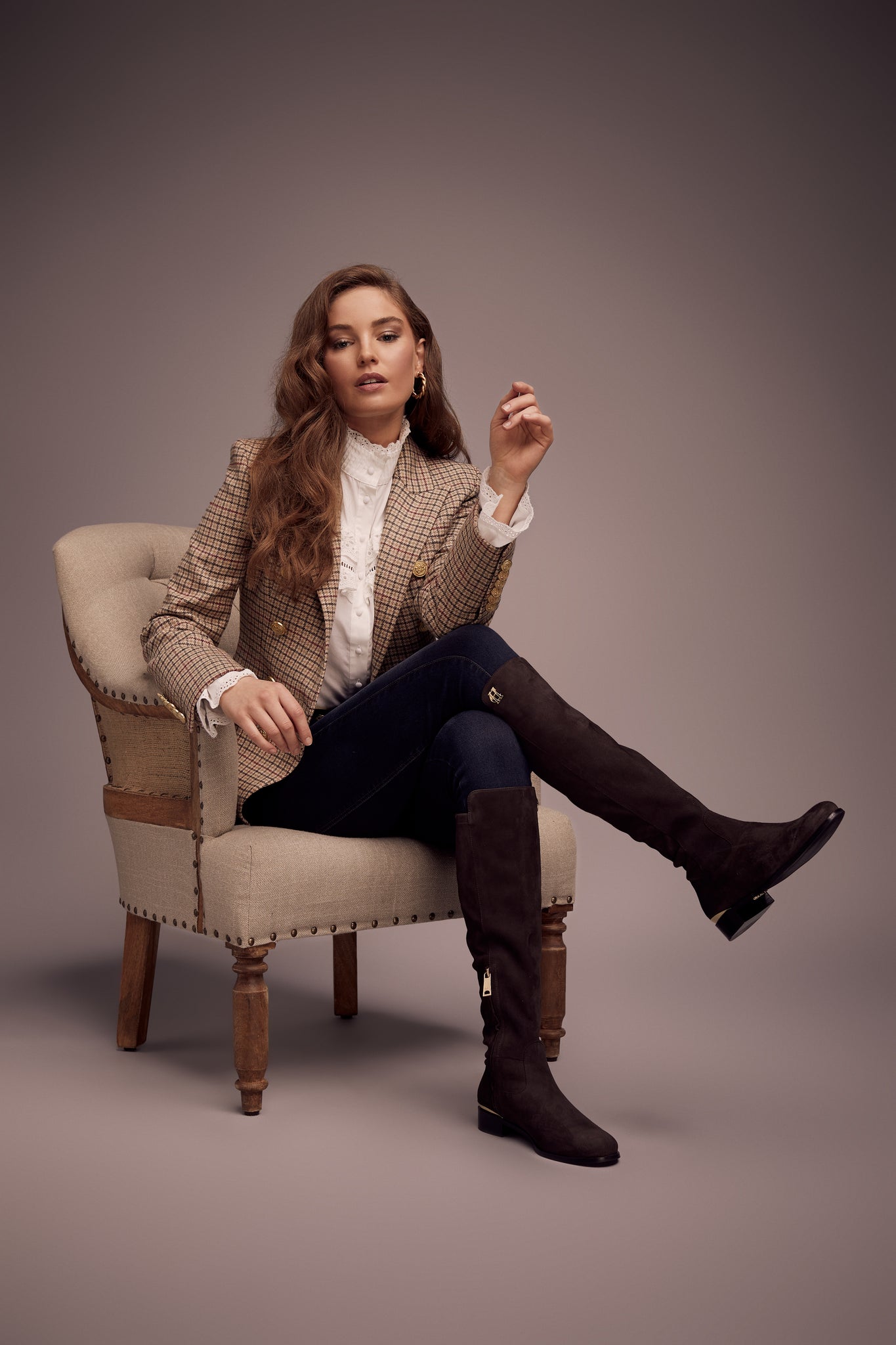 model sat on chair wearing British made double breasted blazer that fastens with a single button hole to create a more form fitting silhouette with two pockets and gold button detailing this blazer is made from camel black and red check charlton tweed