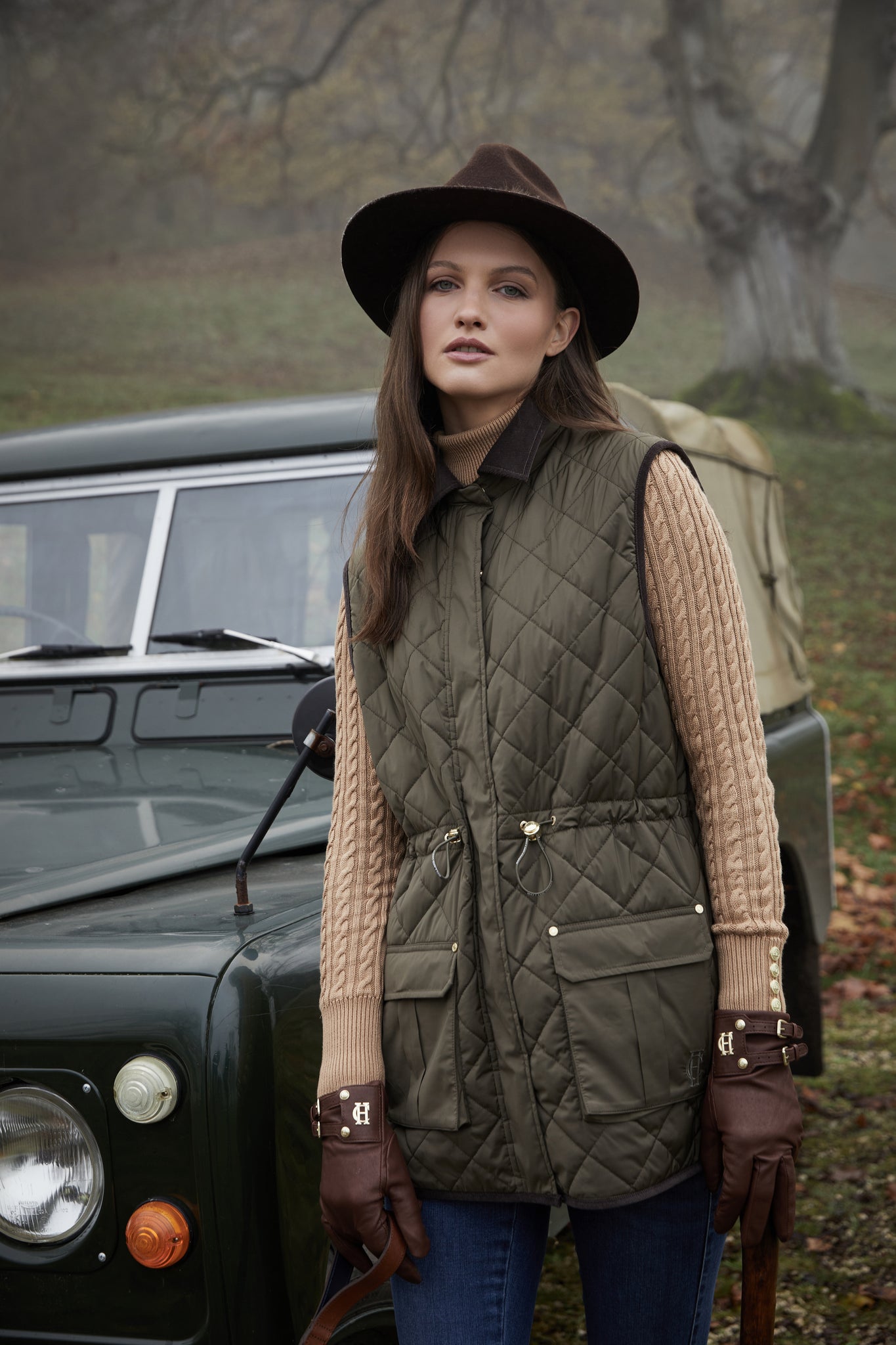 womens khaki quilted gilet with a black collar and seams along the arm holes with drawstring toggles on the waist and two front pockets worn with a beige knitted jumper a dark brown hat and gloves and indigo skinny jeans