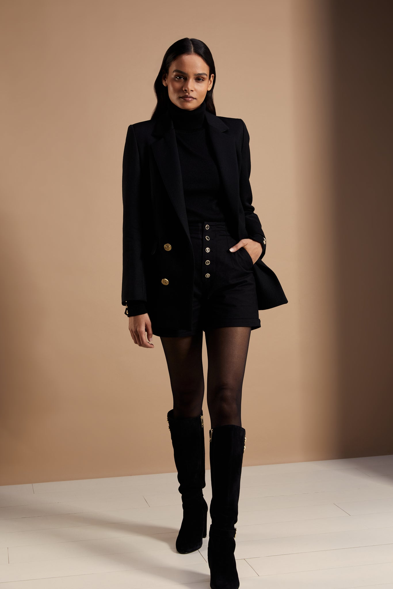 womens black tailored shorts with two single knife pleats and black and gold front button detail with a turned up hem worn with black roll neck and black double breasted blazer