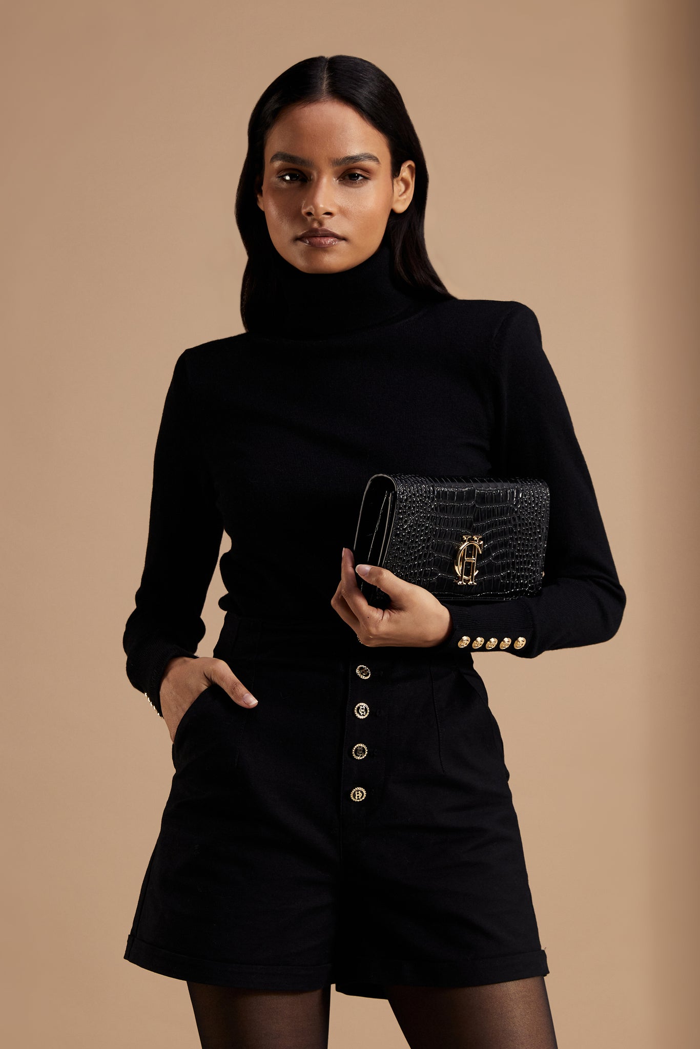 womens black tailored shorts with two single knife pleats and black and gold front button detail with a turned up hem worn with black roll neck and black croc embossed clutch bag