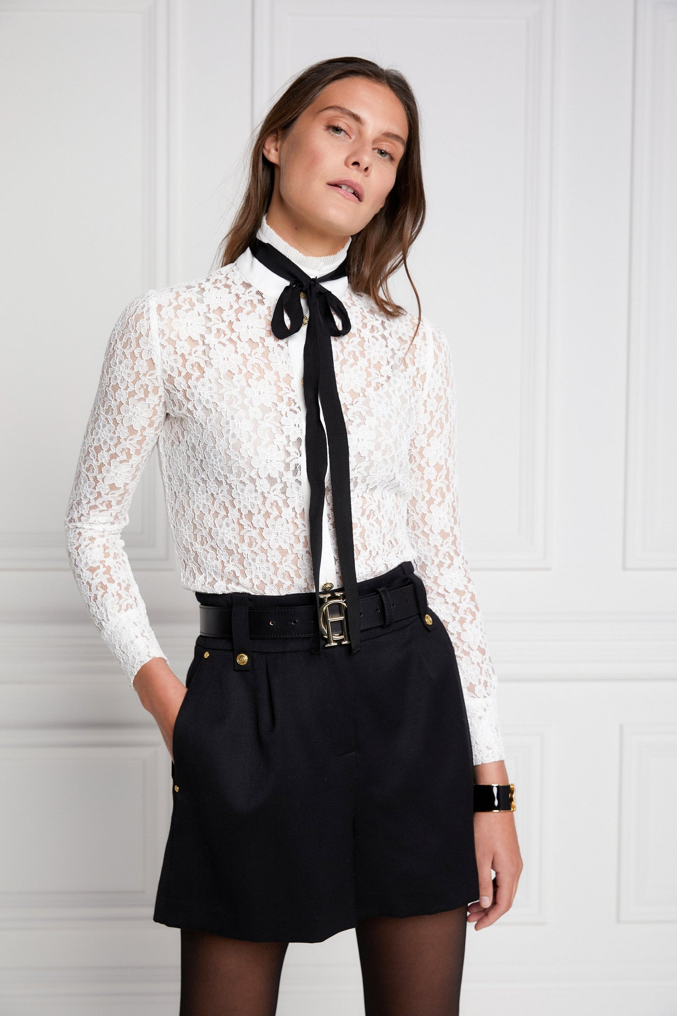 womens black high rise tailored shorts with two single knife pleats and centre front zip fly fastening with twin branded gold stud buttons and side hip pockets with branded rivet detailing at top and bottom of pockets worn with white lace shirt with black tie around collar